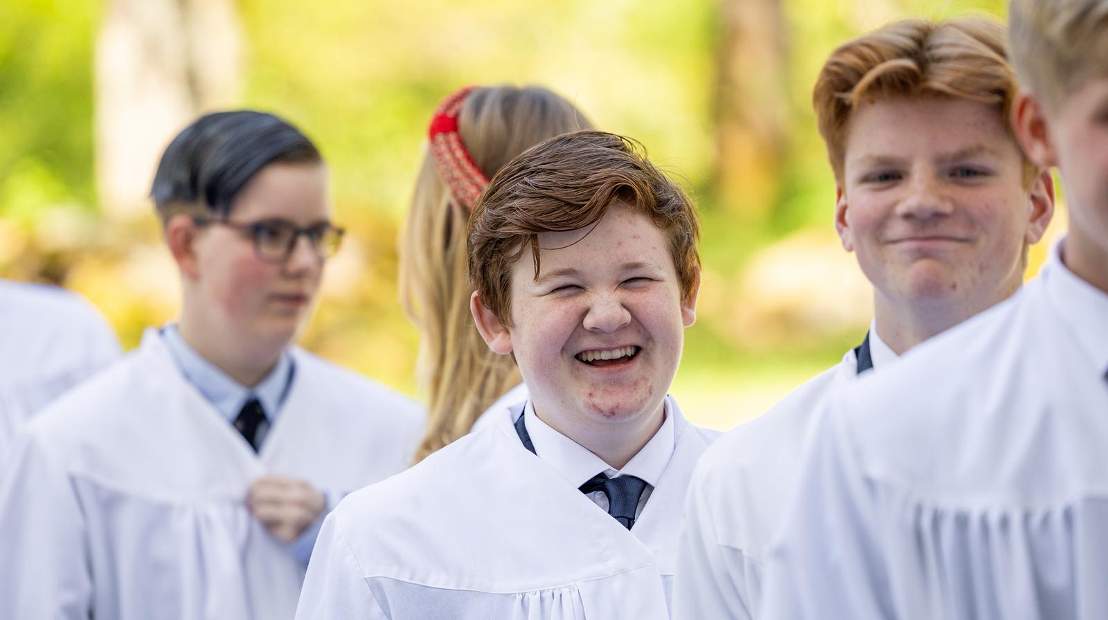 Mange ungdommer i hvite kapper klare for konfirmasjon, en av de smiler s&aring; bredt at &oslash;ynene lukker seg