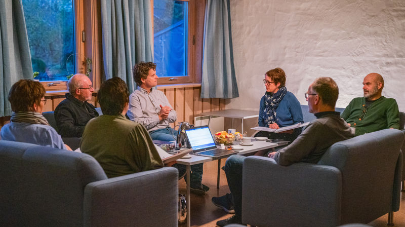 Sju mennesker sitter i sofaer og lenestoler rundt et bord og fiskuterer, pc og sakspapir og kaffekanner på bordet.