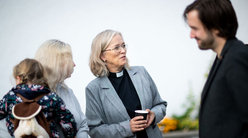 Prest i samtale med kirkegjengere utendørs. Presten har på seg presteskjorte, og har en kopp kaffe i hånda. Alle smiler.