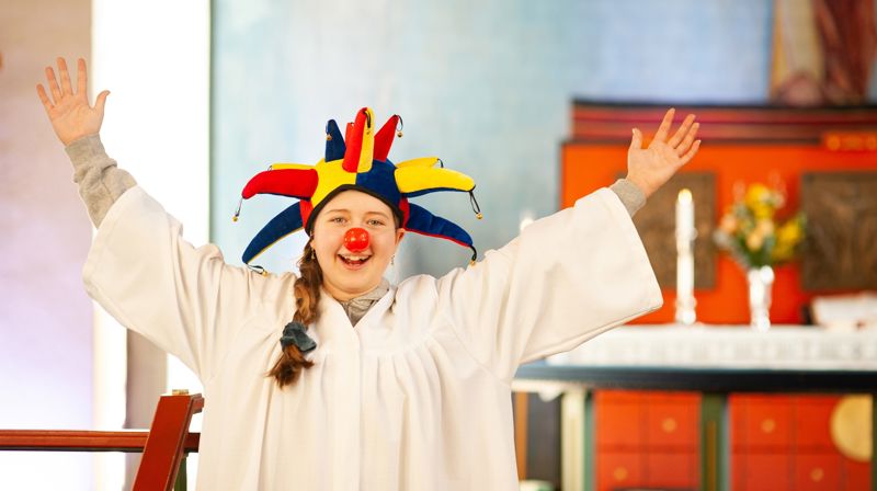 Jente i konfirmantkappe med rød klovnenese og narr-lue på hodet