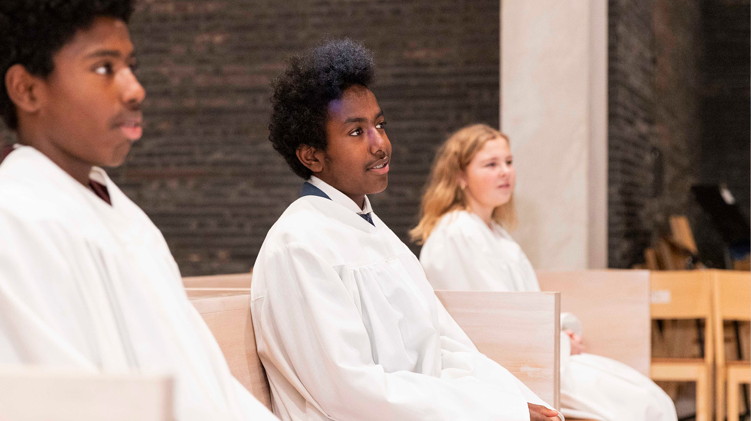 To gutter og en jente sitter på en kirkebenk i konfirmantkapper. 