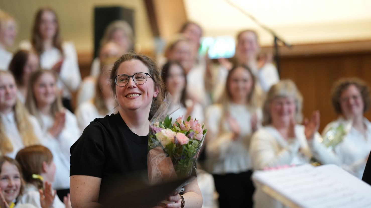 Smilende dirigent med blomsterbukett foran kor kledd i hvitt. Foto.