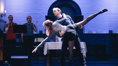 To ungdommer som danser på scenen i en kirke, med forsangere i bakgrunnen