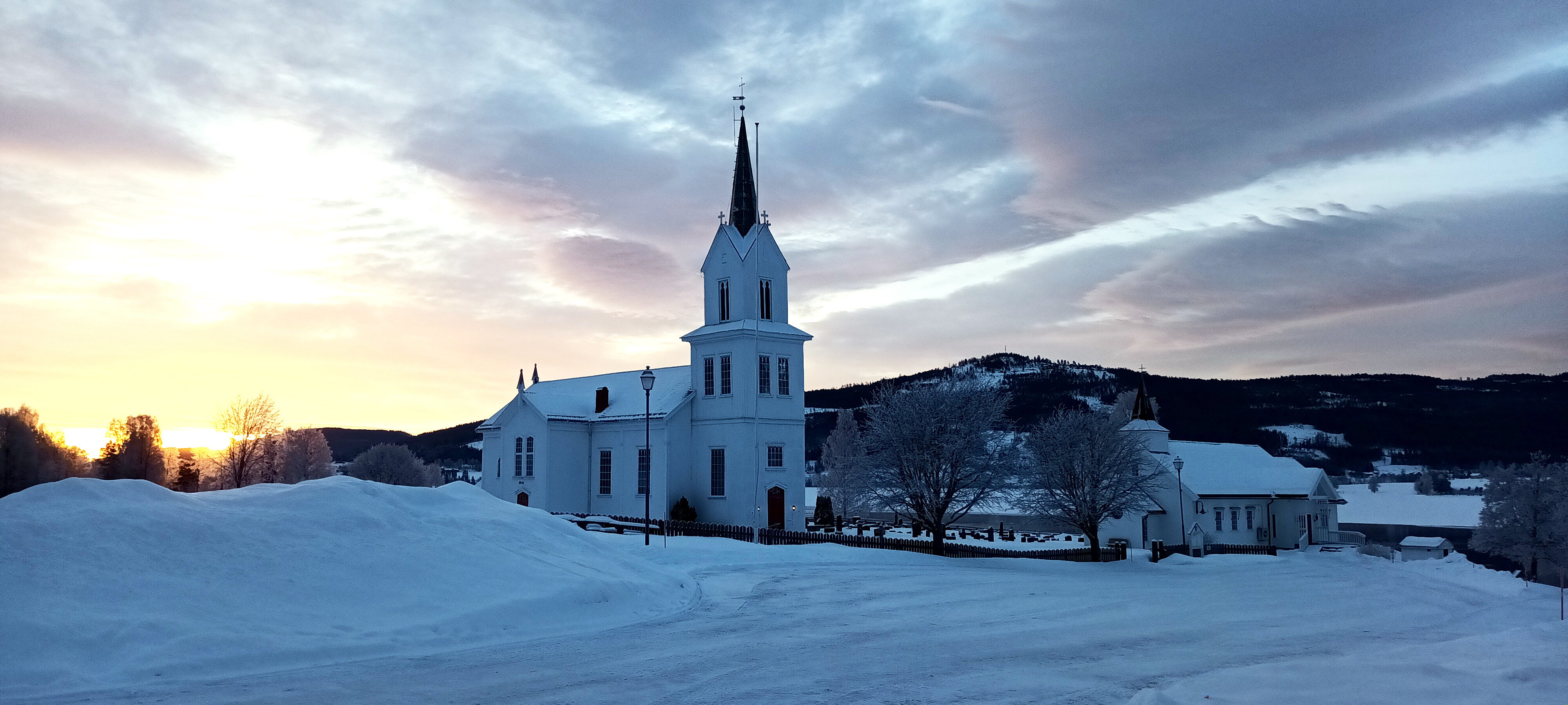 Olberg kirke og kirkestua dekket av snø mot en bakgrunn med skyer og sola som skinner gjennom.
