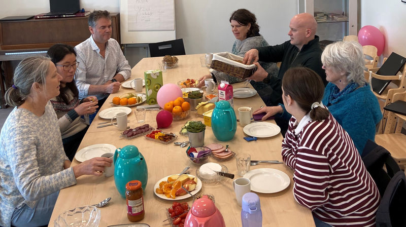 Sju menneske sitt på eit langbord og snakkar og et. Det er frukt og pålegg på bordet.