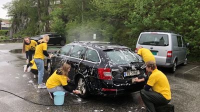 Fleire ungdommar i gule t-skjorter vasker ein bil på ein parkeringsplass