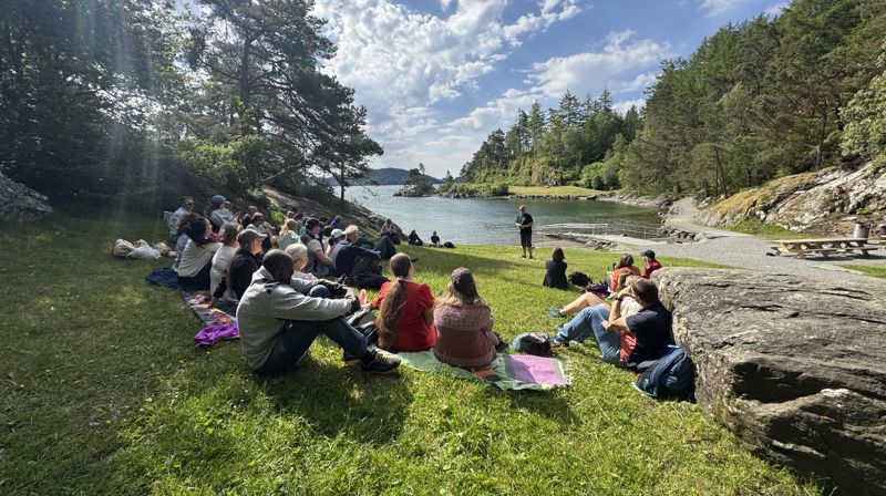 Mennesker sitter i en halvsirkel ute på gresset ved en sjø, en mann står fremme og snakker. det er sol ute