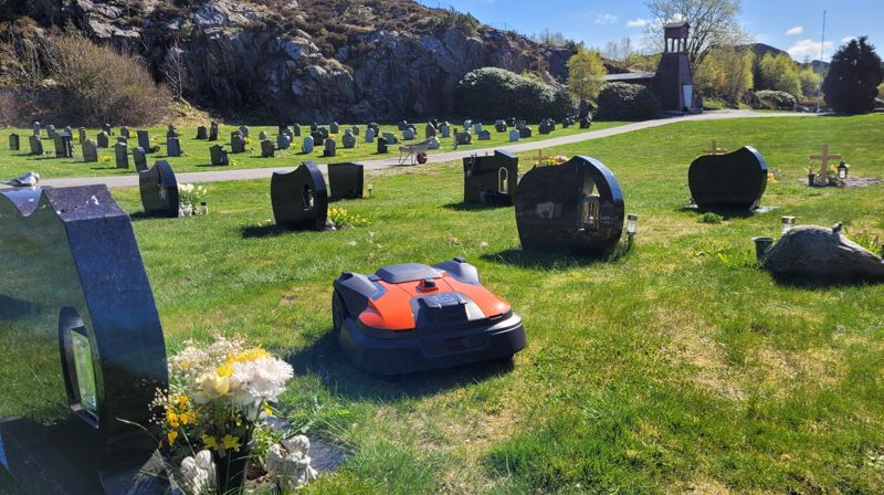En robotgressklipper går på gresset mellom gravstøtter, det er sol, i bakgrunnen ser vi et brunt bygg.