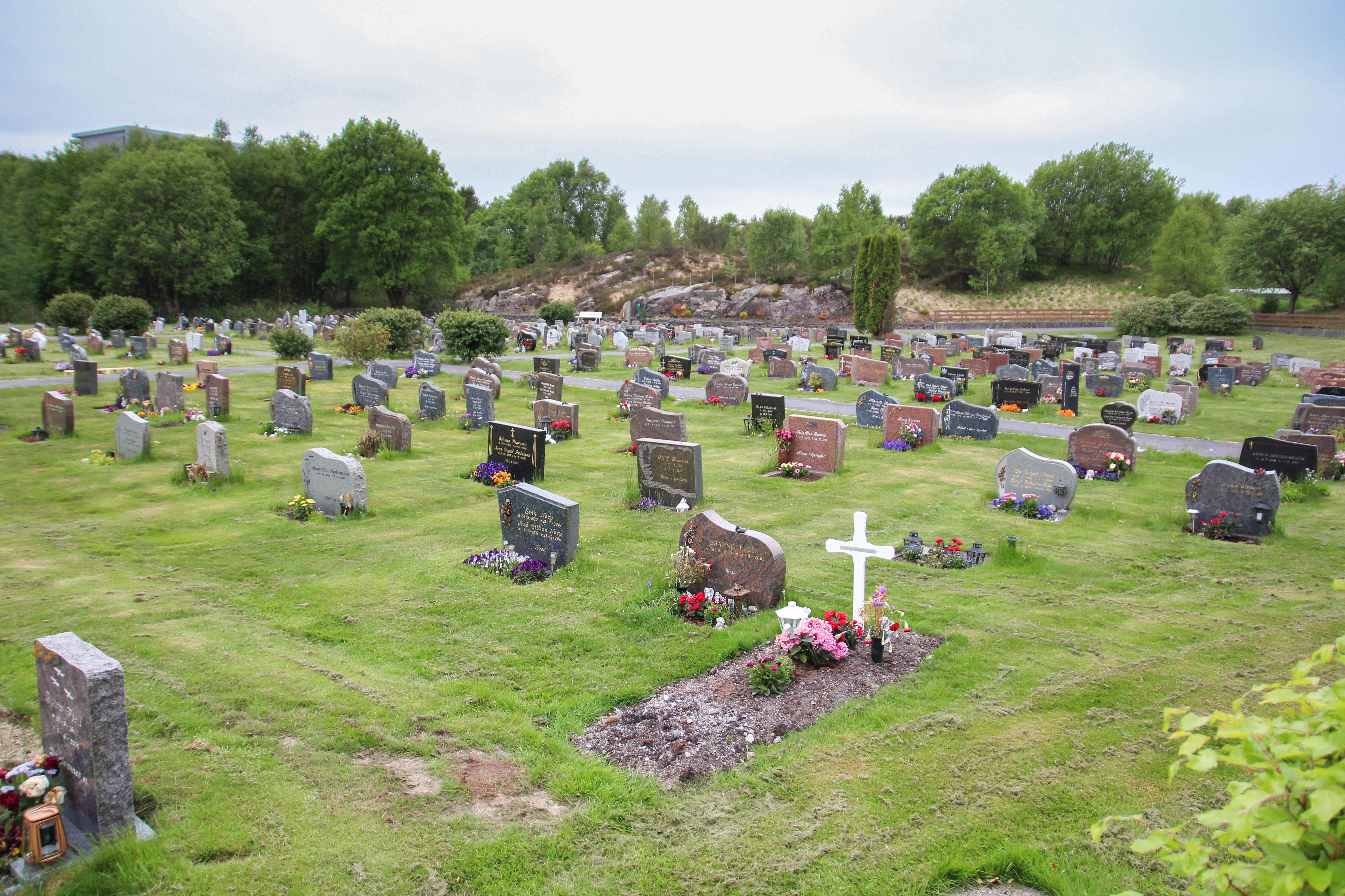 Oversikt over Straume gravplass med en relativt ny grav fremst, den har femdeles jord foran seg og roserbukett plasset oppå. Et hvit kors med navnet på bak rosene. 