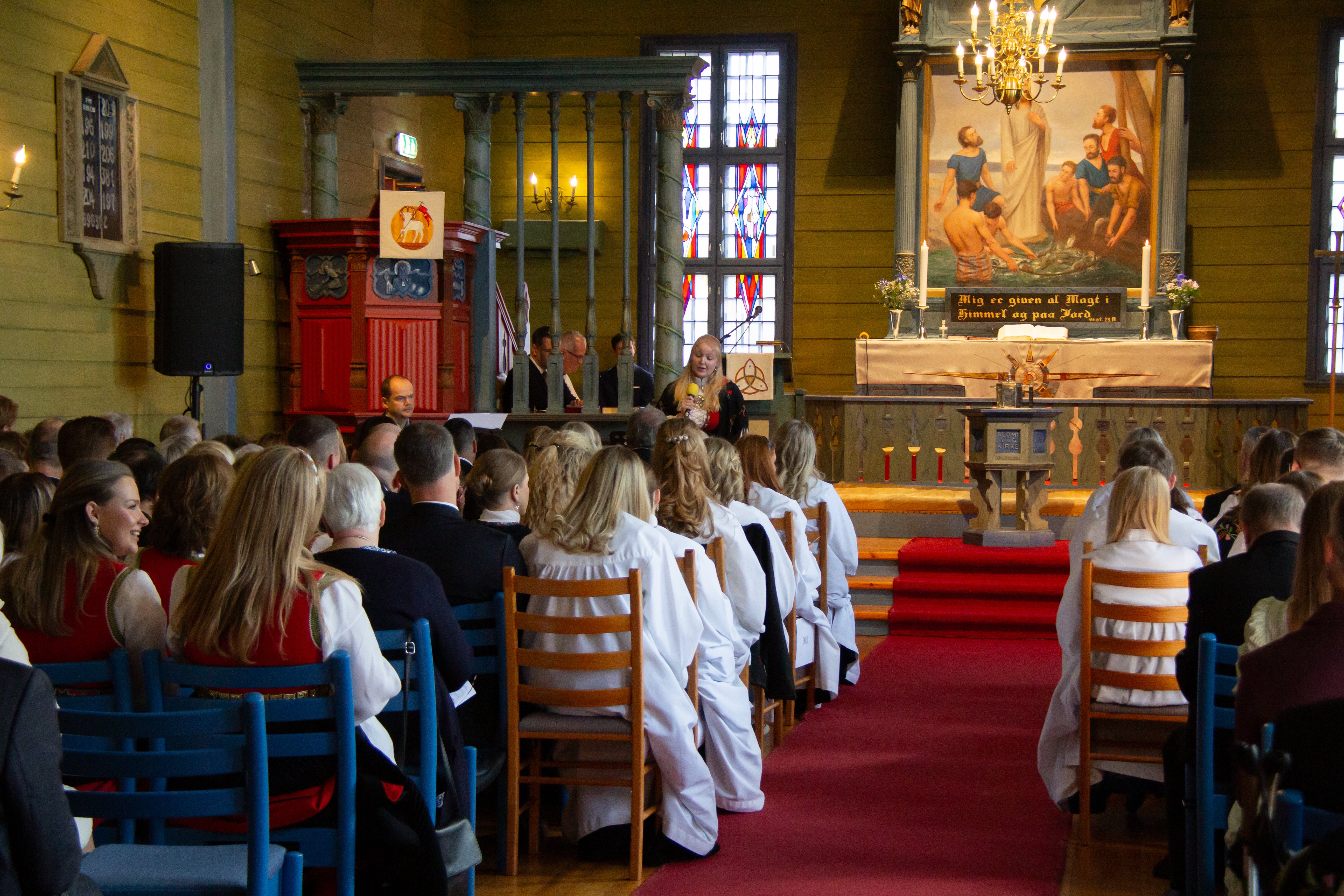 Inne i blomvåg kyrkje sitter konfirmantar i hvite kapper på stoler langs midtgangen, kirken er fylt opp med familie og venner i festklær