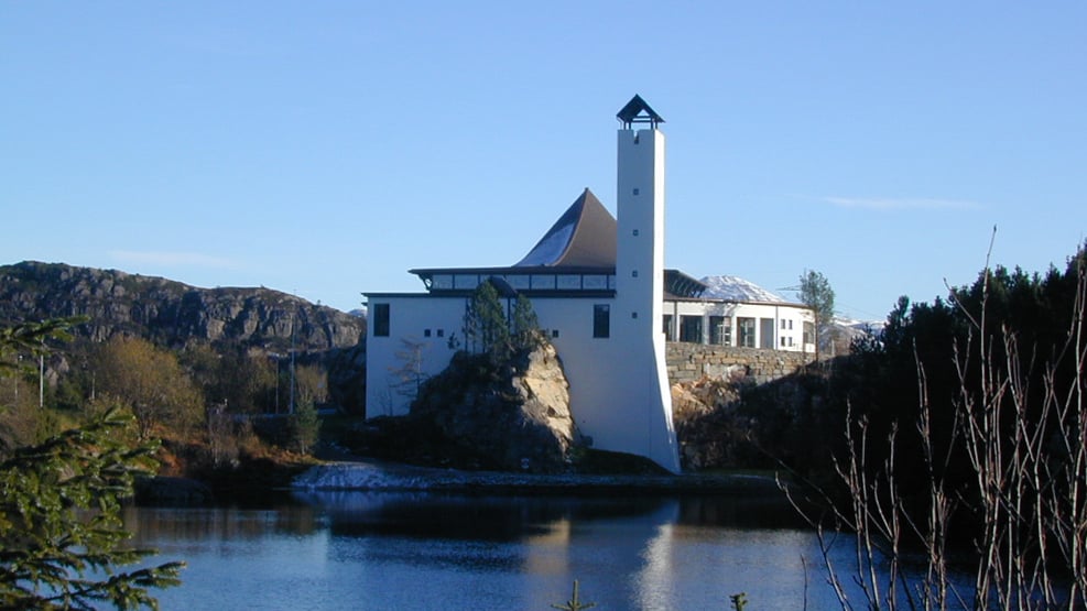 Foldnes kyrkje med vannet i forgrunnen. Det er blå himmel og snø på fjellet i bakgrunnen.  