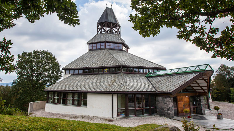 Sund kyrkje med grå og blå himmel i bakgrunnen og grønne trær som rammer inn kyrkja.