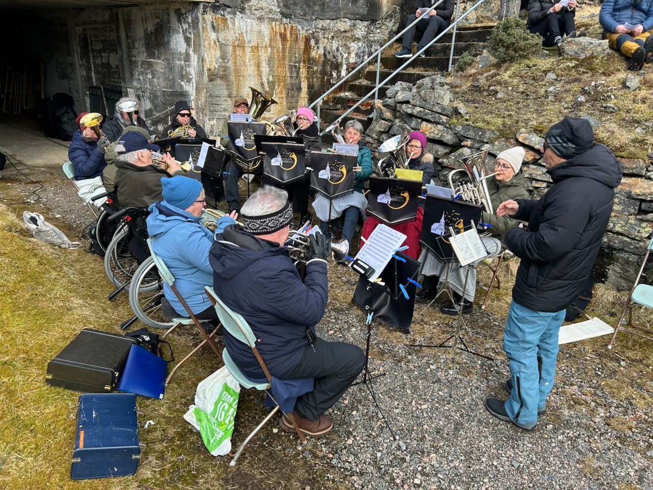 Et brassband sitter p&aring; stoler i en halvsirkel, de spiller ute og er godt kledd