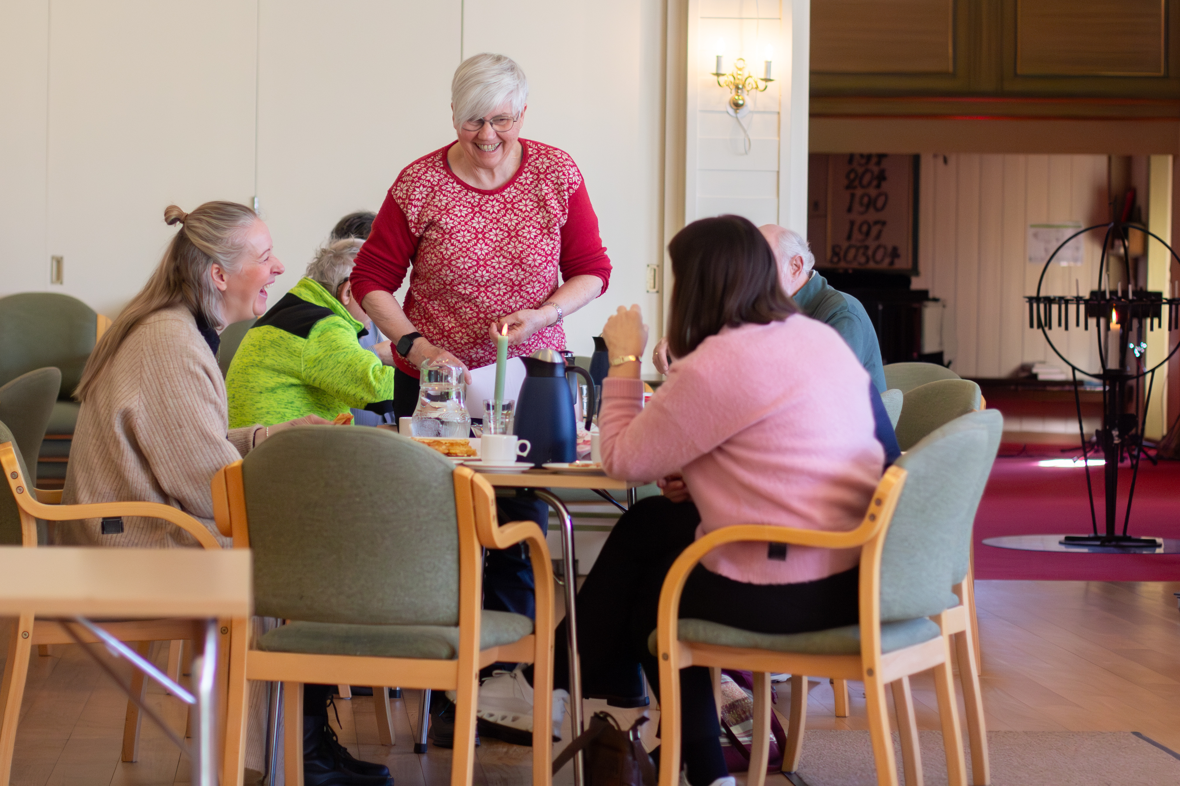 En dame står ved et bord og smiler og ler sammen med andre som sitter ved bordet
