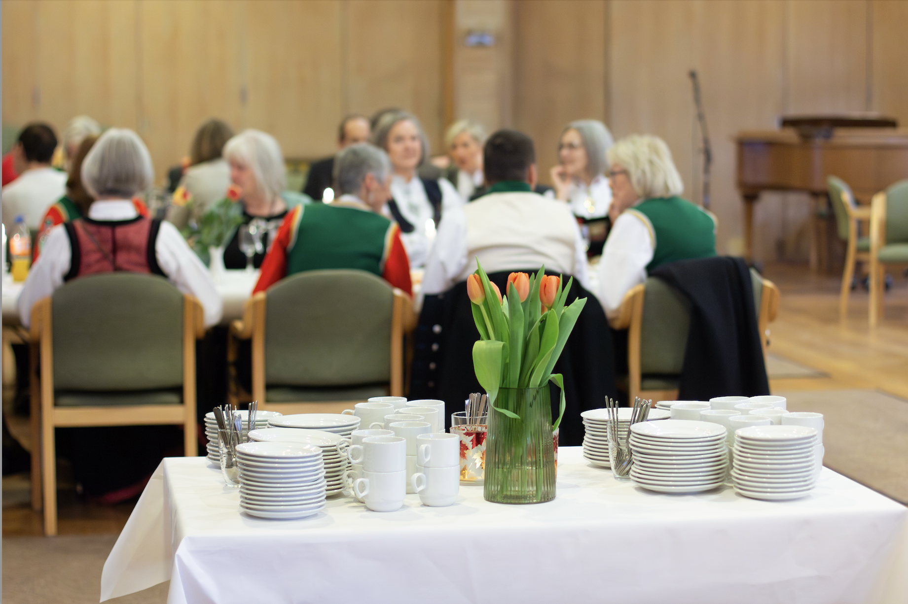 Et bord med hvit duk, tulipaner, kopper, skjeer og fat. Mennesker som i sitter ved bord, mange har bunad.