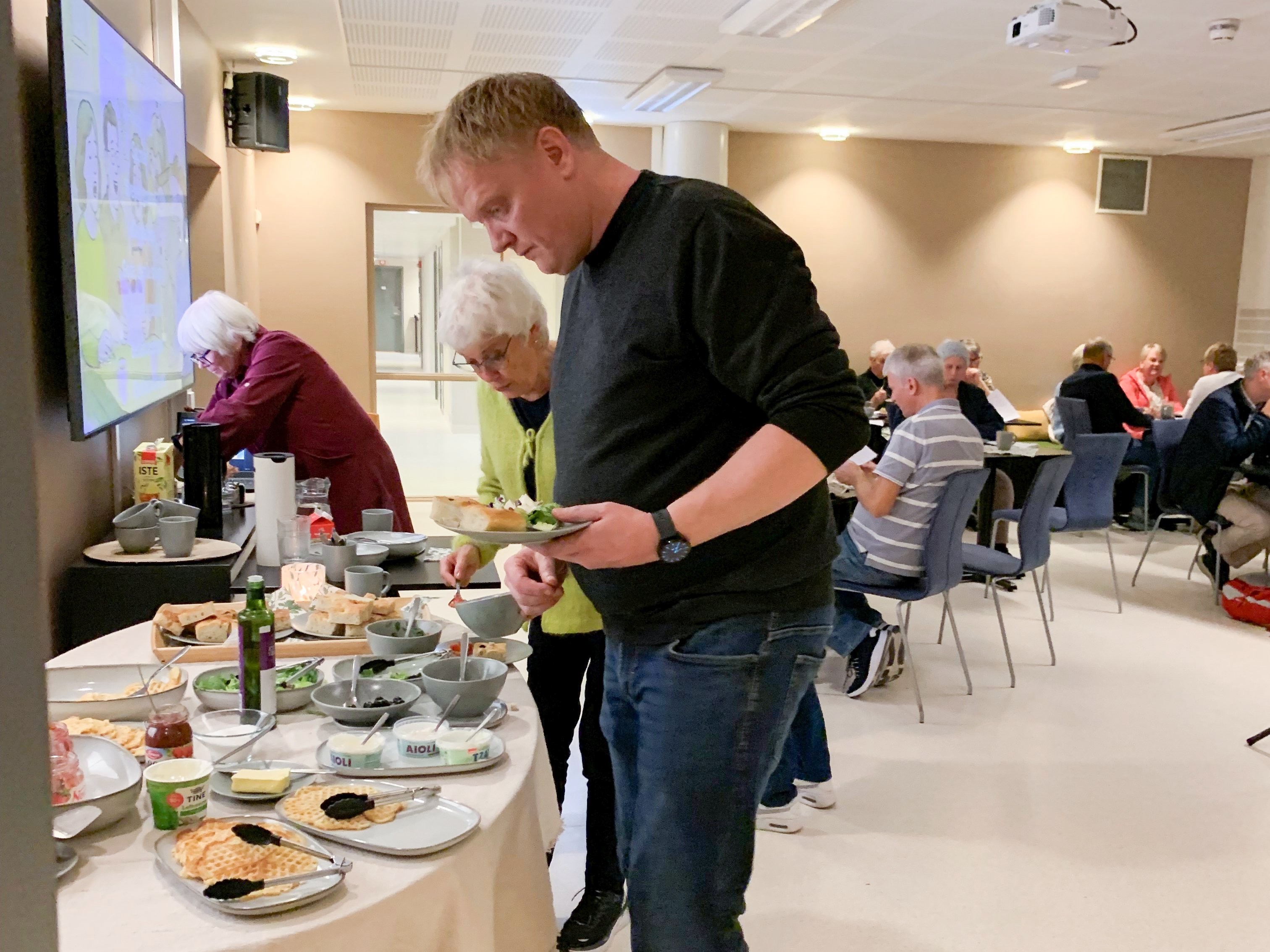 En mann serverer seg fra et bord med ulik mat, vaffler og foccatia. mennesker sitter rundt bord i bakgrunnen