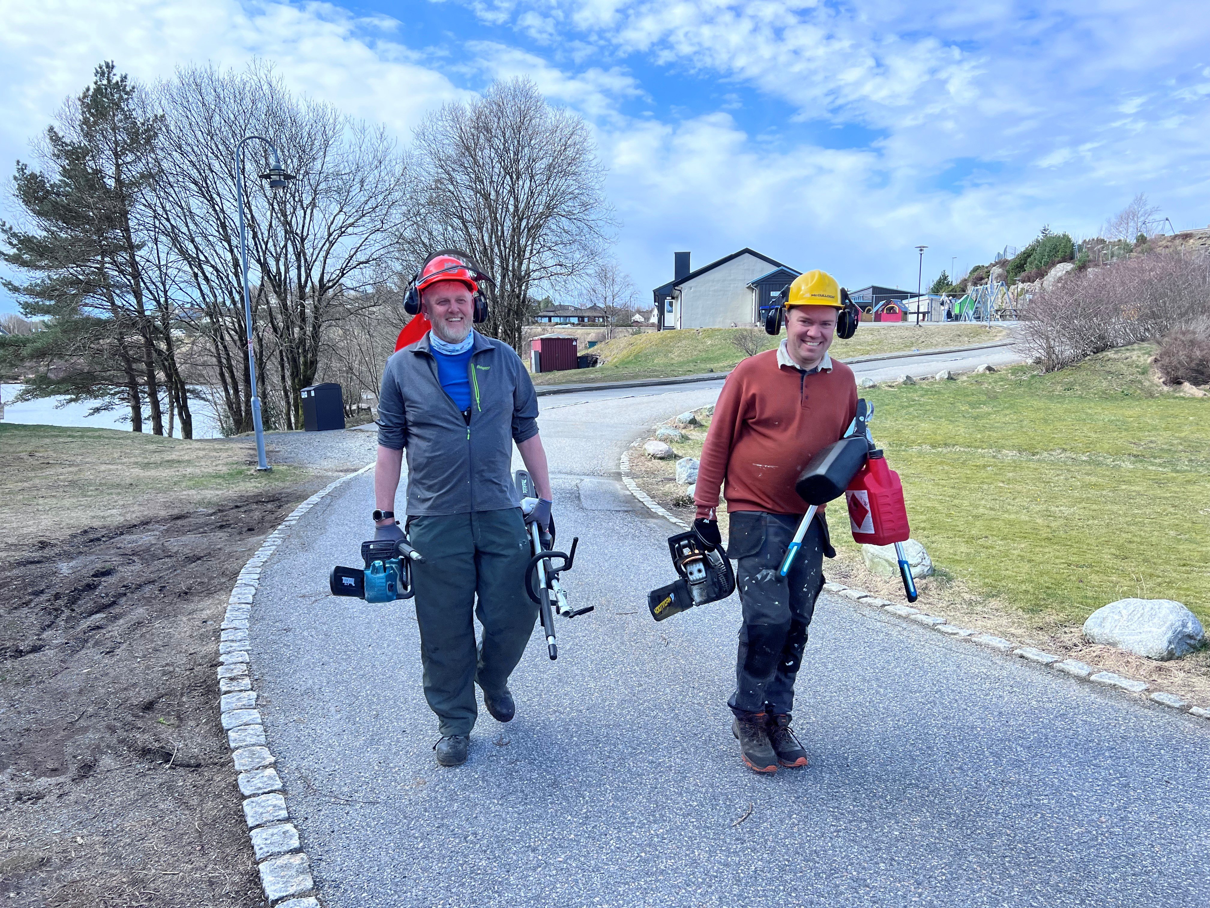 To menn kommer gående smilende, de har hjelm på hodet og verktøy i hendene