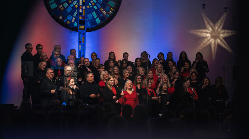 Koret står stilt opp framme i kyrkja, dei er kledd i svart og rødt. ei stjerne reflekteres i eit vindauge
