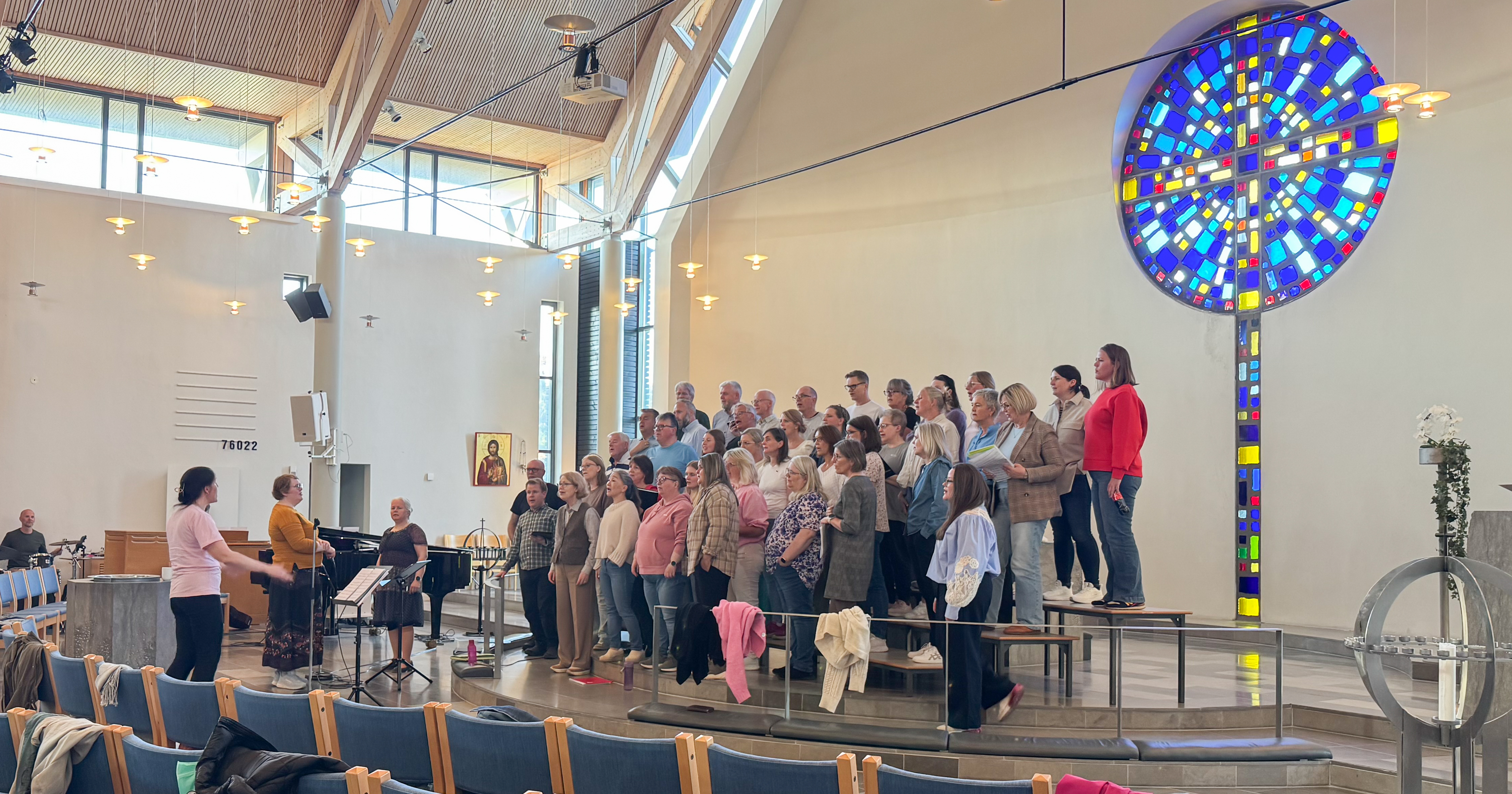 Et kor med rund 50 mennesker står framme bak alterringen og øver, en dirigent står til høyre i bildet sammen med to solister.