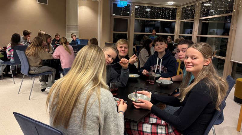 Barn/ungdom sitter på stoler rundt bord og spiser grøt. de nærmeste ser i kameraet og smiler