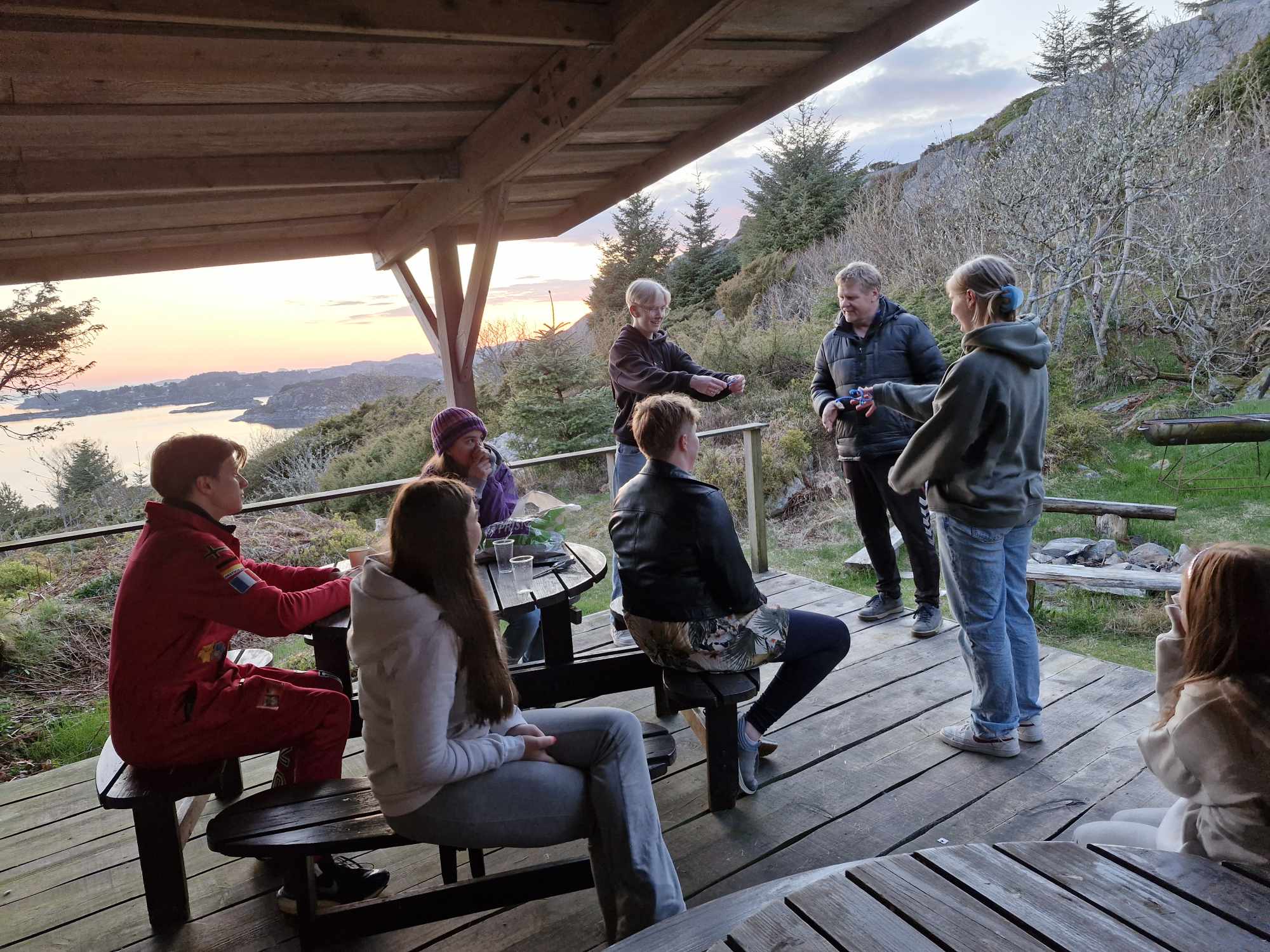 Ungdommar sit ute på ei terasse og hører og ser på en mann som snakkar, det er solnedgang i bakgrunnen