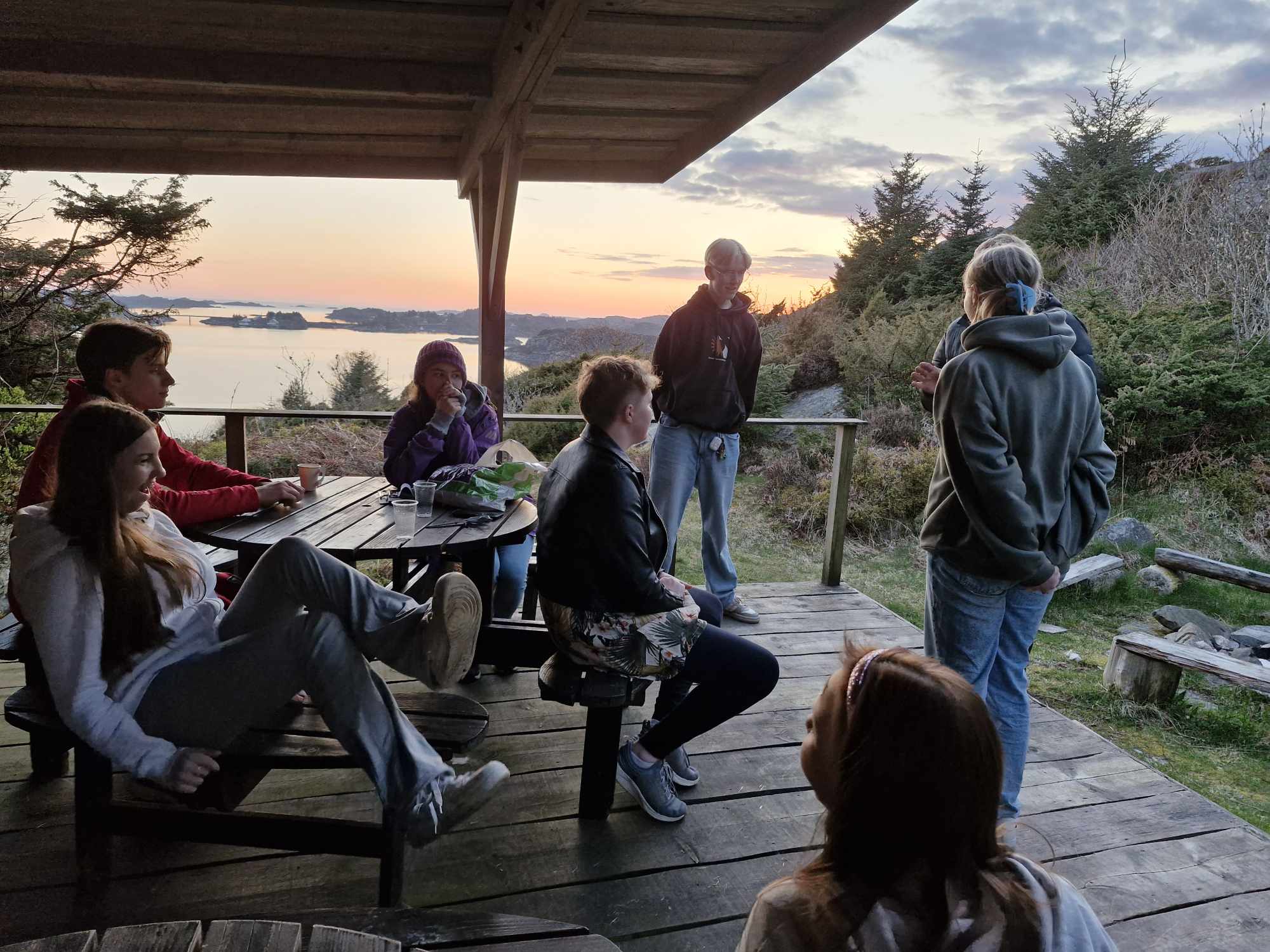 Ungdommar sit ute på ei terasse, det er solnedgang i bakgrunnen