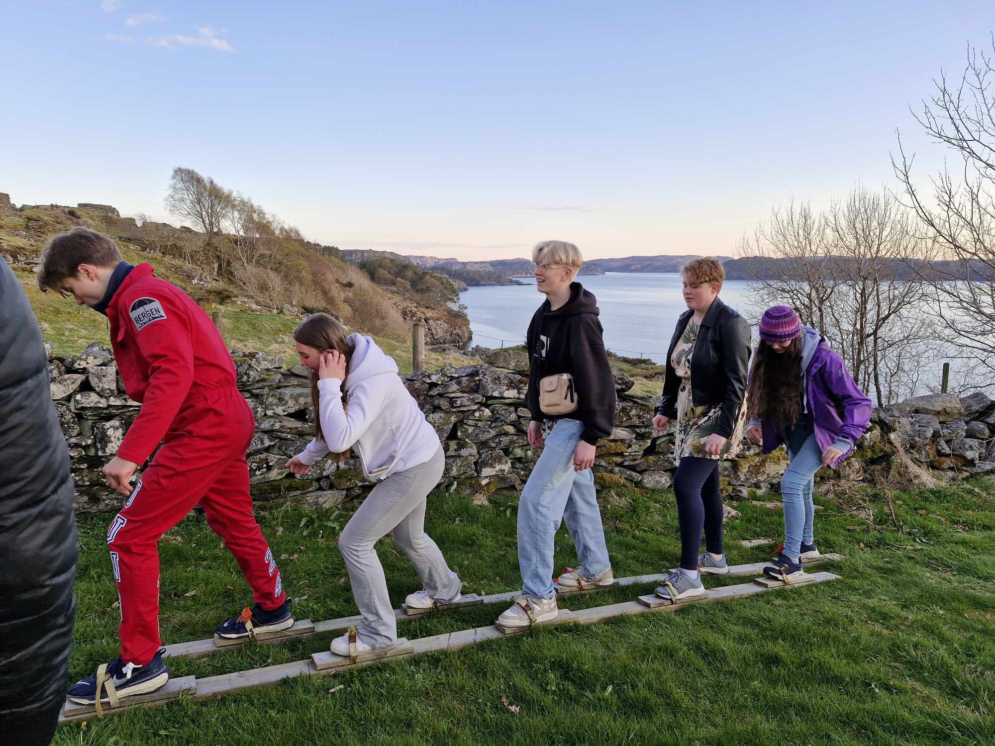 Fem ungdommar står på to plankar, festa på beina som ski, og balanserer bortover graset