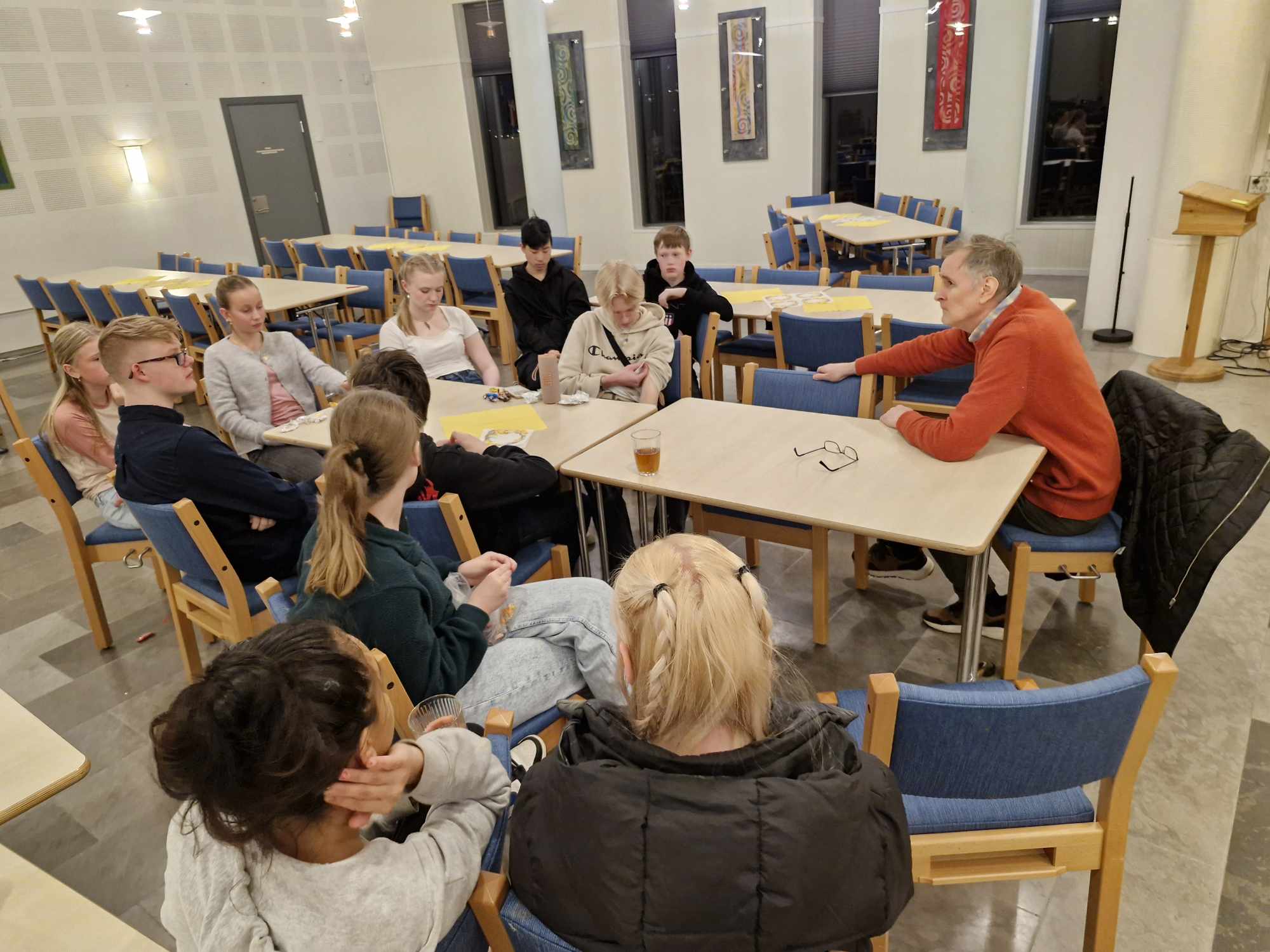 Ungdommar sit rundt eit bord, i enden til høgre sit ein eldre mann og snakkar