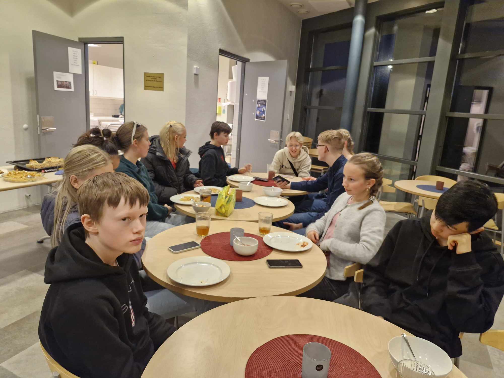Ungdommar sitt rundt små runde bord som er satt saman, nokre pratar og nokre kjedar seg