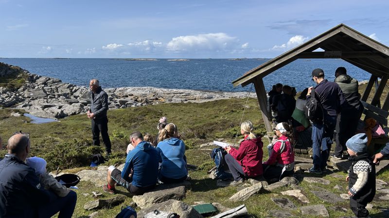 Mennesker står og sitter på en plass rett ved havet, noen sitter også inni en liten benkplass med tak over.