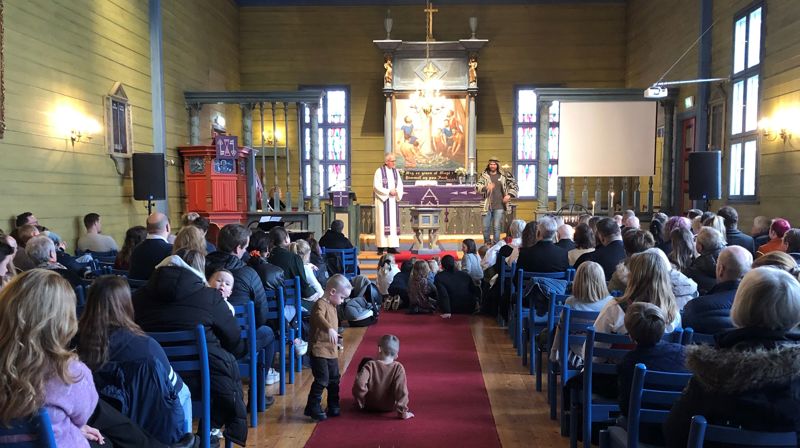 En prest og pedagog står framme i Blomvåg kyrkje, på gulvet framfor dei sit mange barn og høyrer på. Kyrkja er full av menneske som sit på stolane.