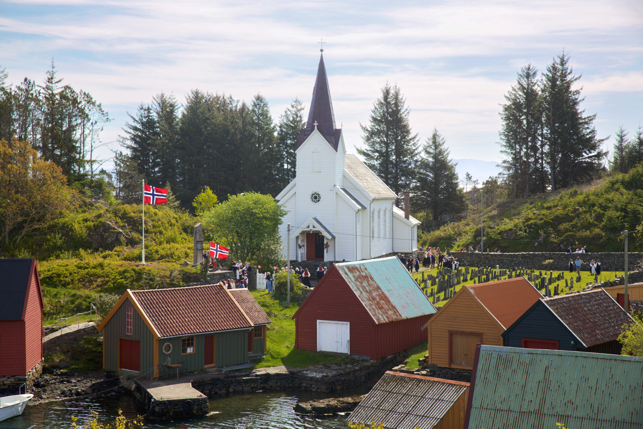 En hvit trekirke med flere naust framfor seg. Flere flagg er heist og man kan skimte mennesker framfor kirken