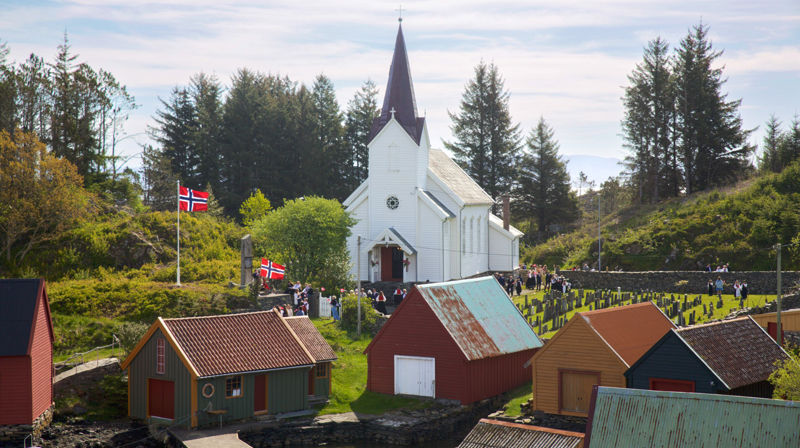 En hvit trekirke med flere naust framfor seg. Flere flagg er heist og man kan skimte mennesker framfor kirken