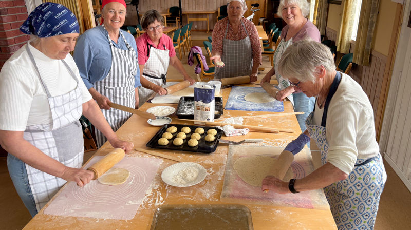 Fflere damer sår rundt et stort bord og baker og ruller ut lefser