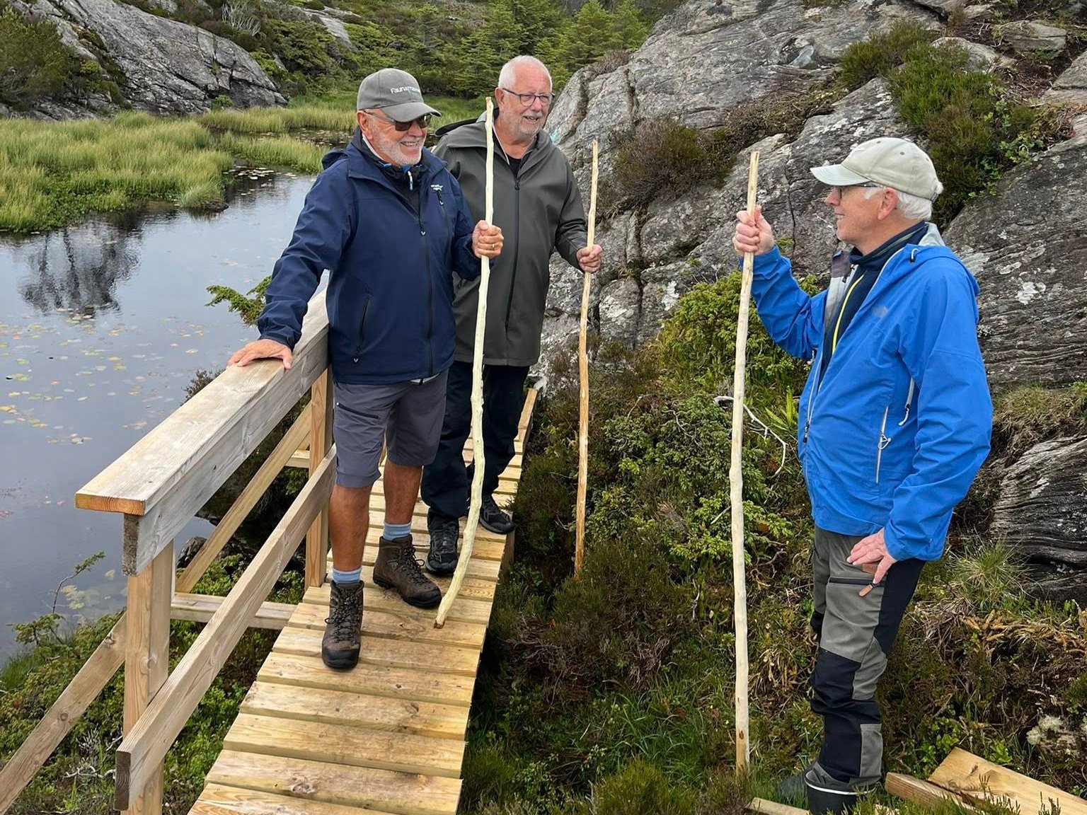 Tre menn står på en tursti med hver sin vandrestav og smiler til hverandre