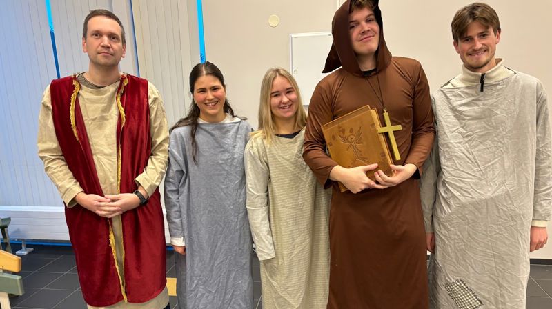 Tre menn og to damer er kledd ut i gammeldagse kostymer, de står på rekke og smiler. En mann holder en stor bok.