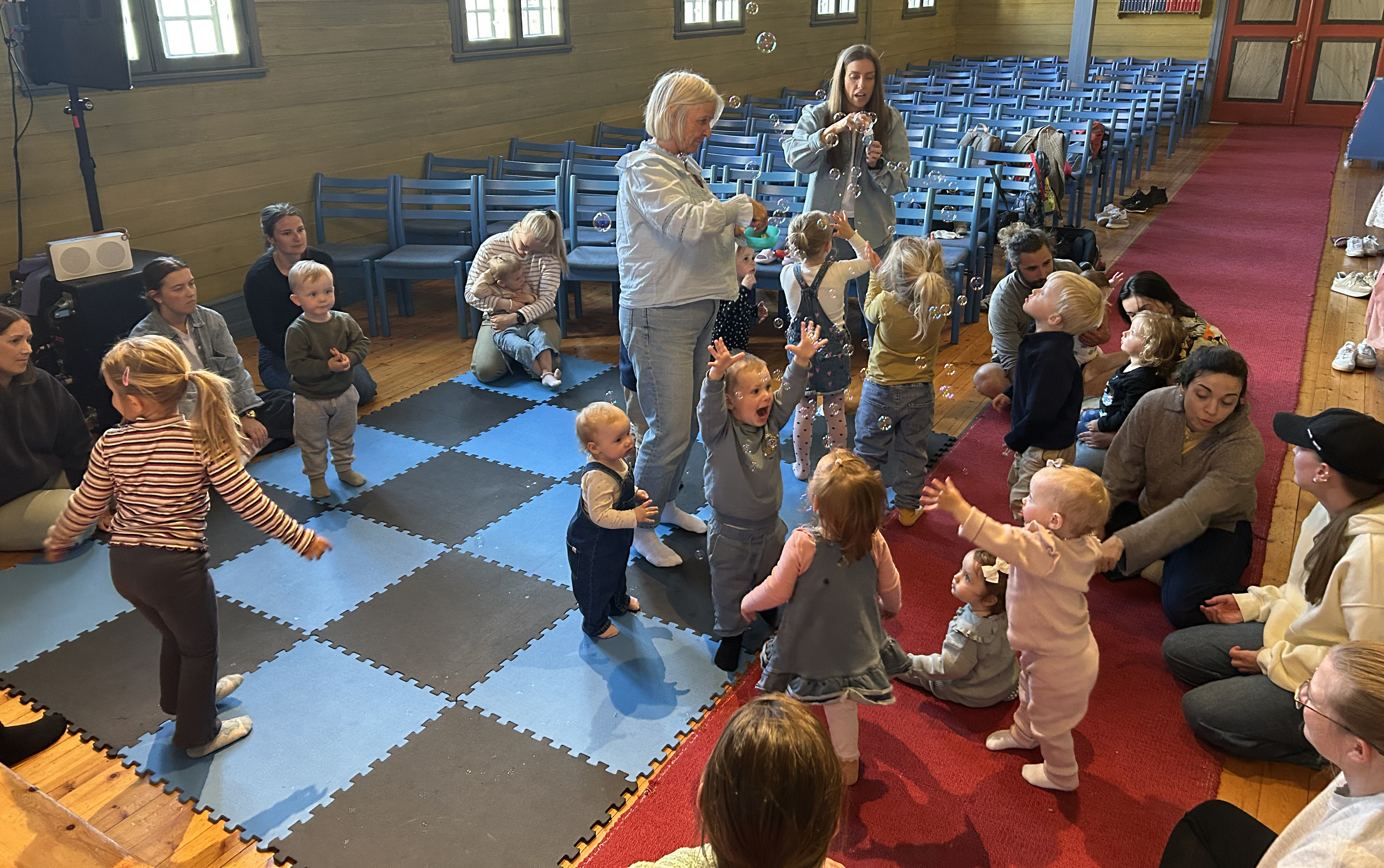 Små barn og voksne står og sitter på matter inni kirken, to damer holder maskiner som blåser ut såpebolber. barna koser seg og prøver å fange boblene