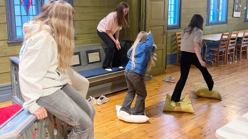Fire tweensjenter, unngår gulvet i en lek. en sitter på alterringen, en står på en benk, og to hopper fra pute til pute på gulvet