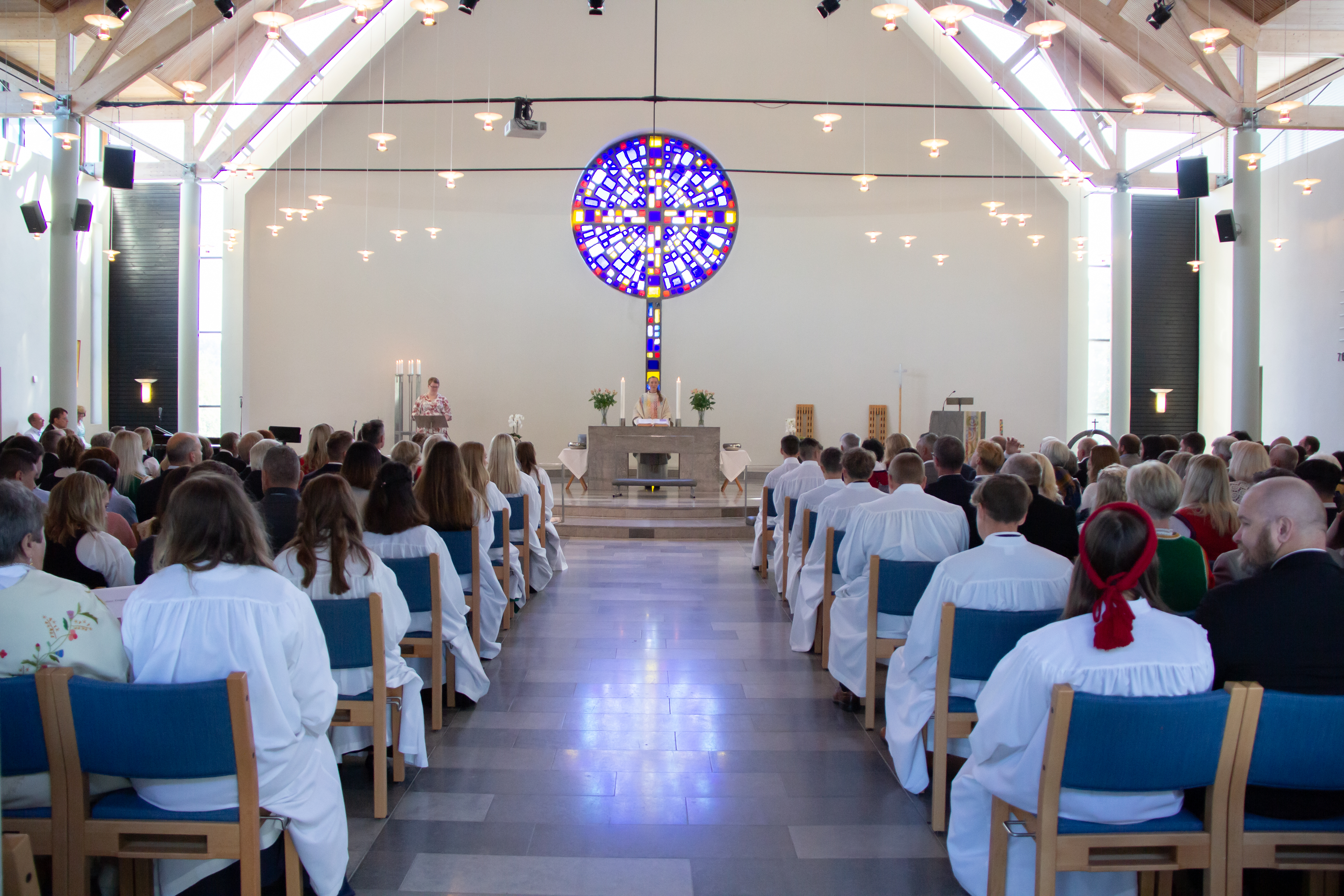 Foldnes kyrkje på ei konfirmasjonsgudsteneste, sett bakfrå. Konfirmantane sit langs midtgongen. Fremst i kyrkja er glasmaleriet: Eit kors i klare fargar.