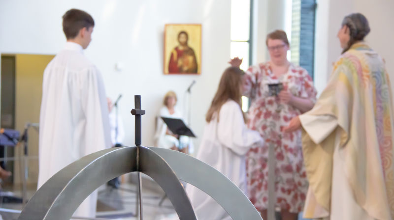 Fokus på kors over lysglobe, uklart i bakgrunnen blir en konfirmant bedt for av konfirmantlærar og prest