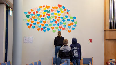 Tre ungdommer står og ser på mange fargerike papirhjerter som henger på veggen