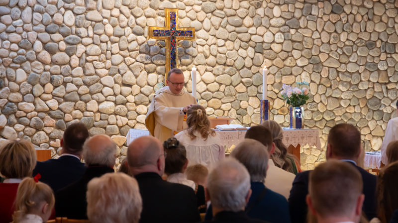 Ein prest ber for ein ungdom på konfirmasjonsdagen. I bakgrunnen: Ein vegg med rullesteinar og eit kors i gull og blått.