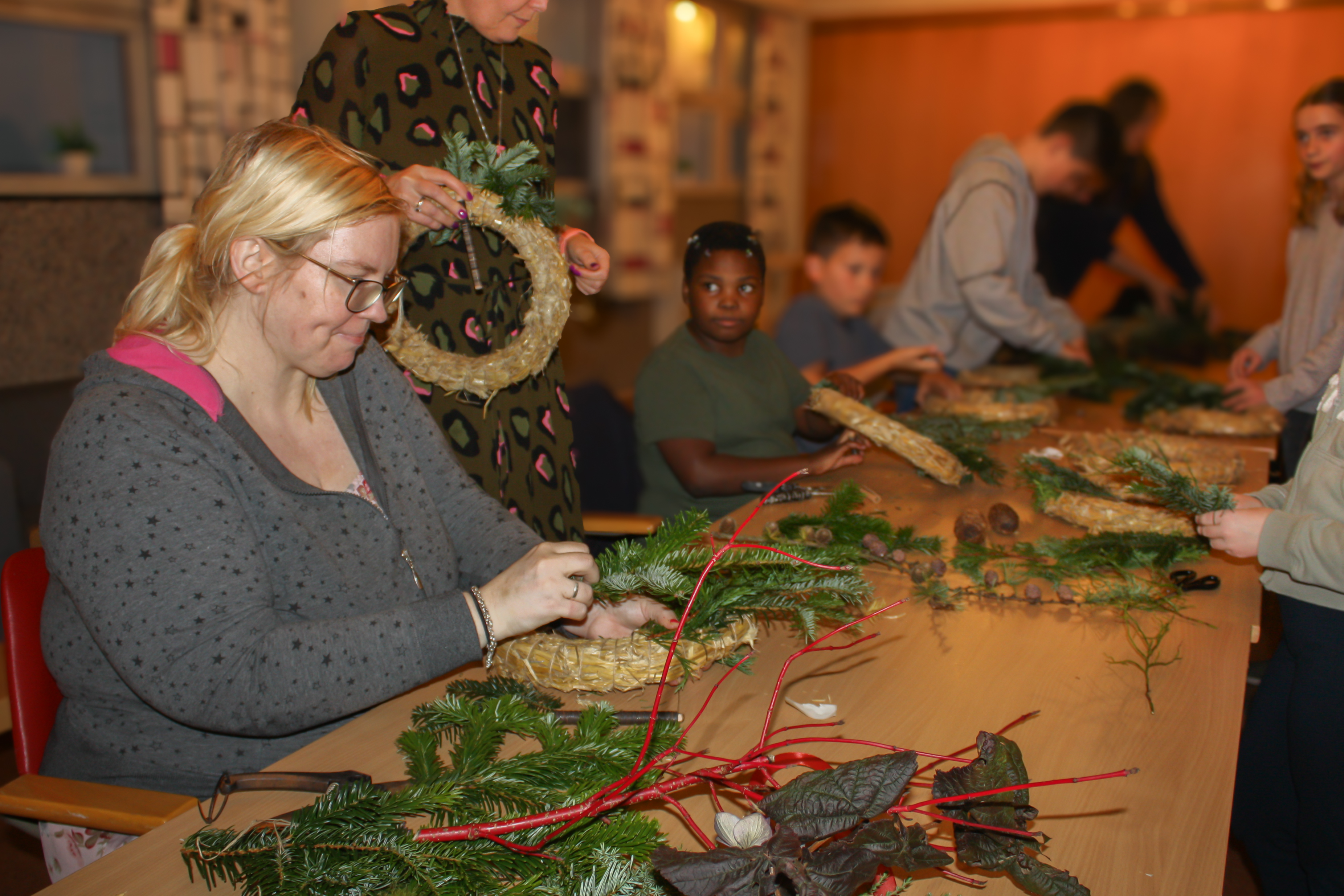 Langs et bord sitter voksne og barn og lager kranser med granbar og pynt