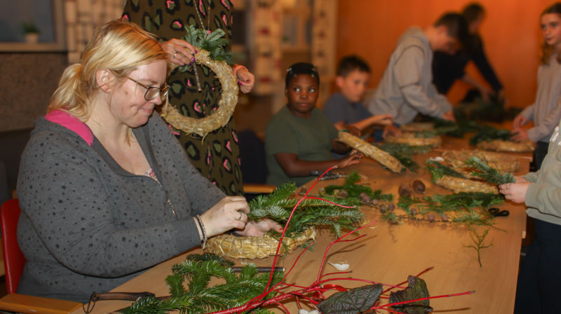Langs et bord sitter voksne og barn og lager kranser med granbar og pynt