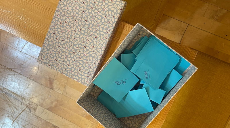 En eske med blomstermønster på en benk, lokket ligger ved siden av og oppi esken ligger brettede, turkise lapper der det står "glede" skrevet på