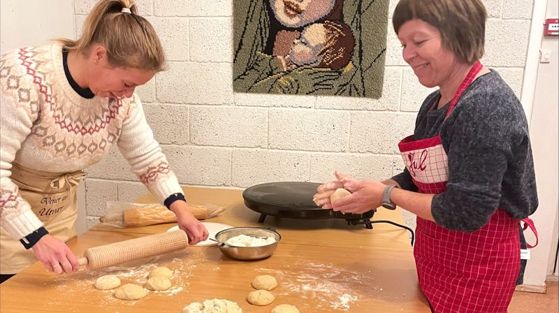 To damer står ved et bord og former og kjevler ut lefsedeig