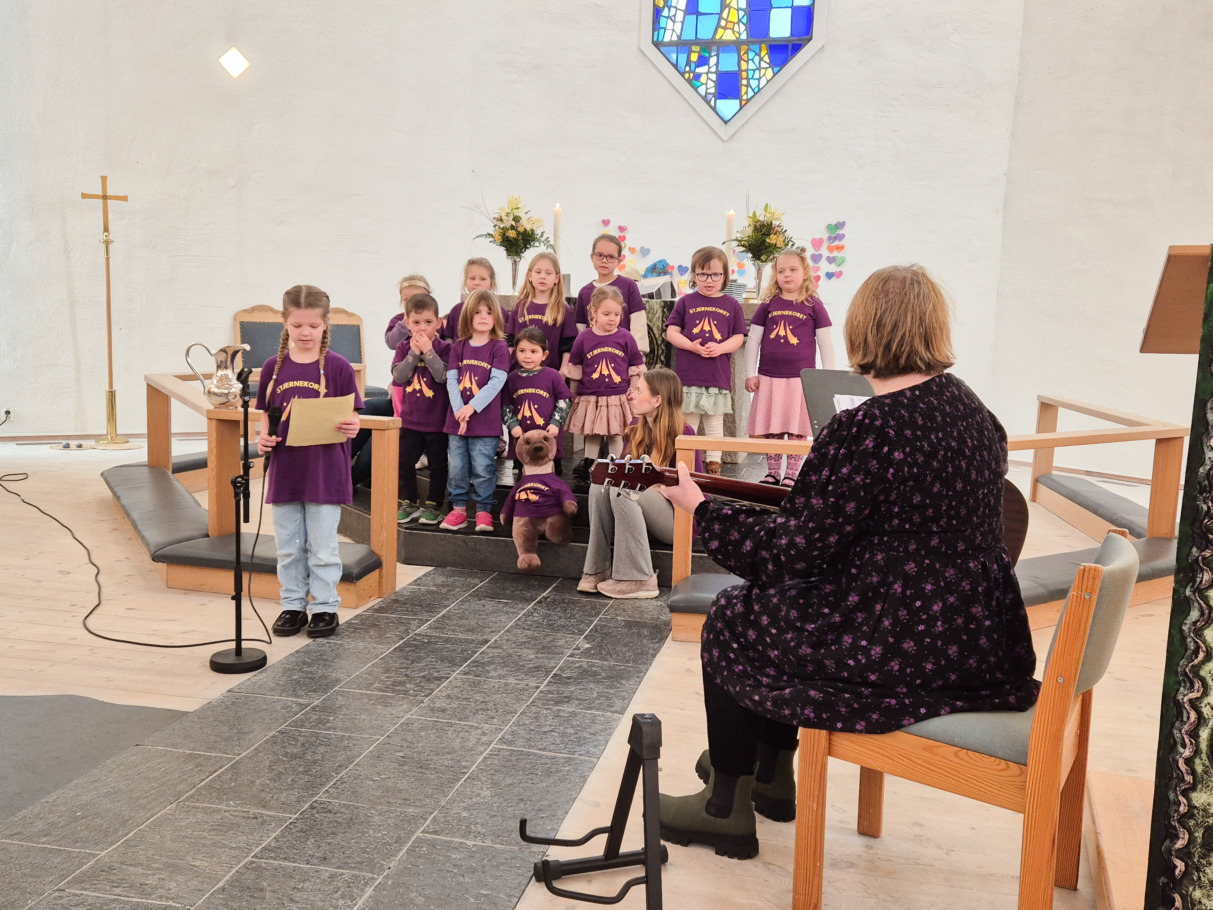 Små barn står fremme i en kirke og synger, de har på seg lilla t-skjorter. En dame sitter på fremste stol og spiller gitar. En jente står fremst og synger i en mikrofon.