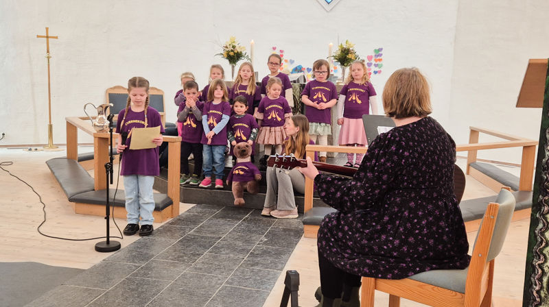 Små barn står fremme i en kirke og synger, de har på seg lilla t-skjorter. En dame sitter på fremste stol og spiller gitar. En jente står fremst og synger i en mikrofon.