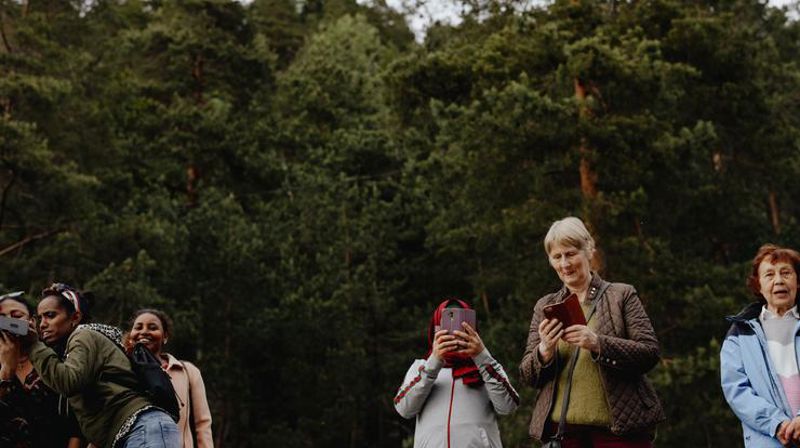 Flere damer står ute i naturen, det er skog i bakgrunnen. Damene har en telefon i hendene, noen tar bilde og noen ser på telefonen.