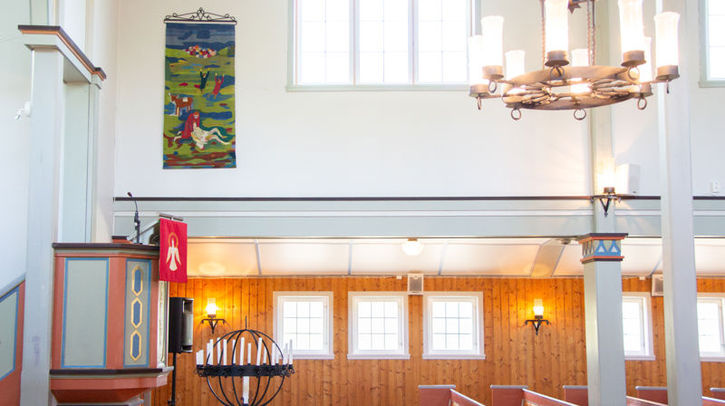 På ei kvit vegg i øvre halvdel av langveggen i kyrkja heng eit veva bilete med grønne og blå fargar